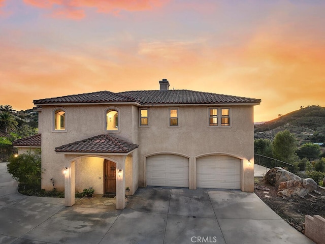 mediterranean / spanish home featuring a tiled roof, concrete driveway, stucco siding, a chimney, and a garage