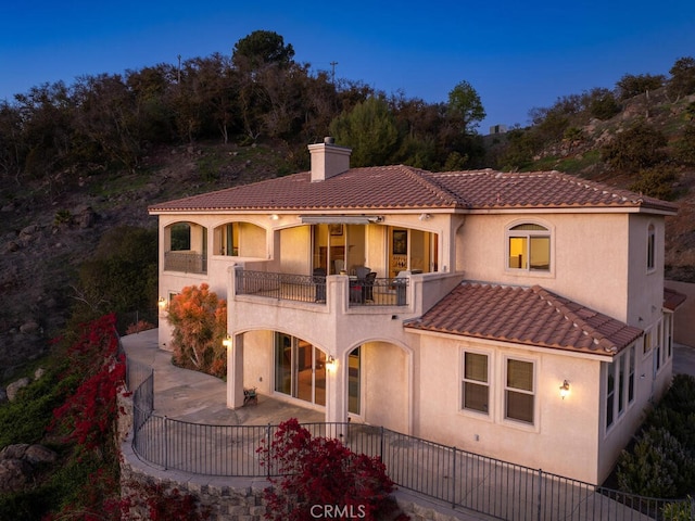 rear view of house with a tiled roof, stucco siding, a chimney, a balcony, and a patio