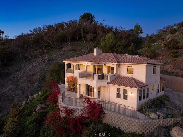 mediterranean / spanish house featuring stucco siding, a tile roof, a balcony, fence private yard, and a chimney