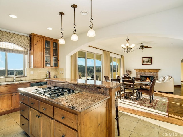 kitchen with a sink, light stone counters, a warm lit fireplace, stainless steel gas stovetop, and brown cabinetry