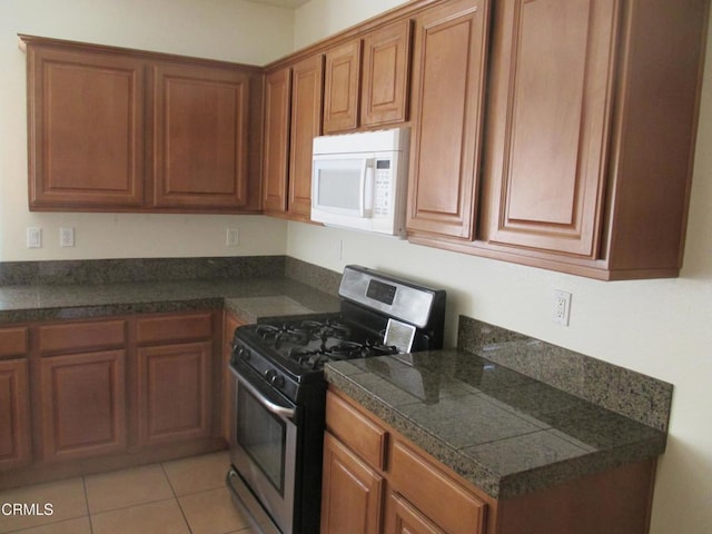 kitchen featuring light tile patterned floors, white microwave, stainless steel gas range, tile counters, and brown cabinets