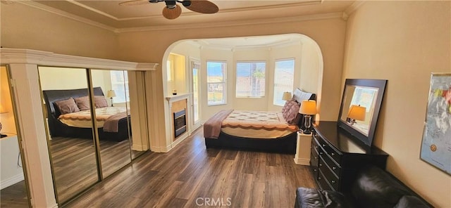 bedroom featuring arched walkways, dark wood finished floors, a glass covered fireplace, a ceiling fan, and crown molding