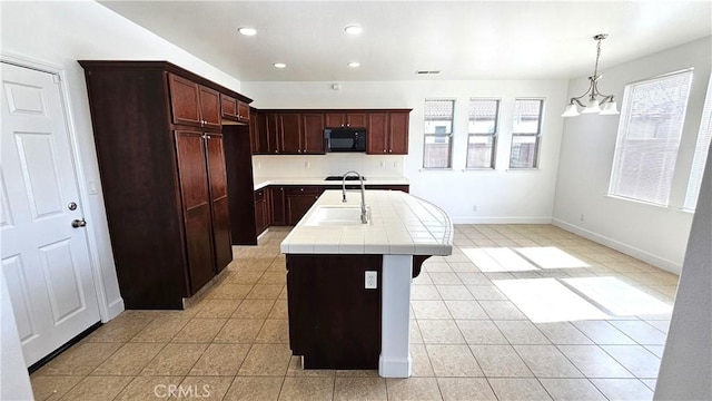 kitchen with black microwave, tile counters, an island with sink, light tile patterned floors, and a sink