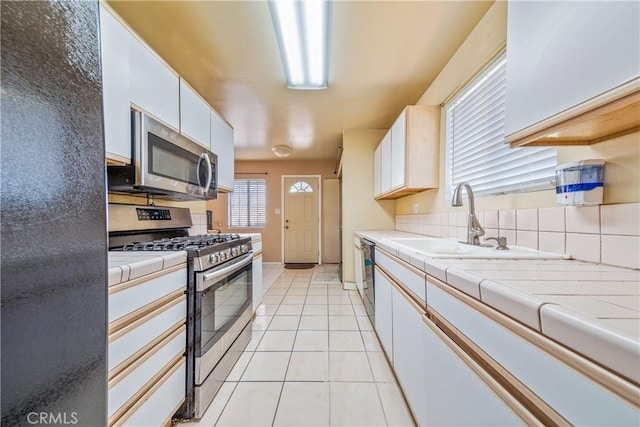 kitchen featuring light tile patterned flooring, a sink, stainless steel appliances, tile counters, and white cabinetry