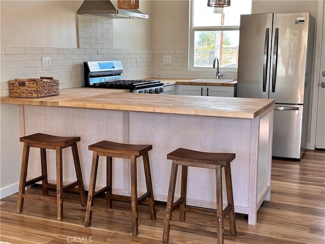 kitchen featuring wood finished floors, a sink, appliances with stainless steel finishes, wall chimney range hood, and butcher block counters