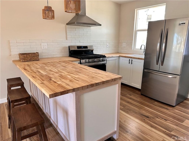 kitchen featuring butcher block counters, a peninsula, stainless steel appliances, wall chimney exhaust hood, and a sink