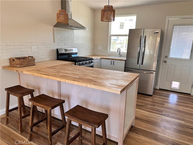 kitchen featuring appliances with stainless steel finishes, a peninsula, butcher block counters, wall chimney range hood, and decorative backsplash