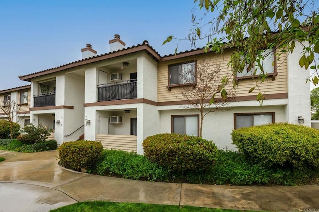 view of front facade with a wall unit AC, a balcony, a chimney, and stucco siding