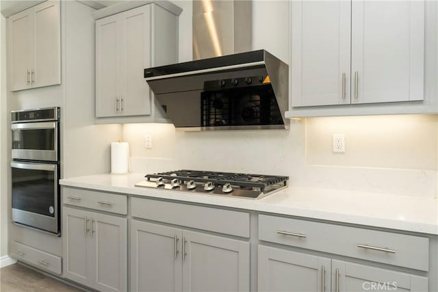 kitchen with gray cabinetry, stainless steel appliances, wall chimney exhaust hood, light countertops, and decorative backsplash