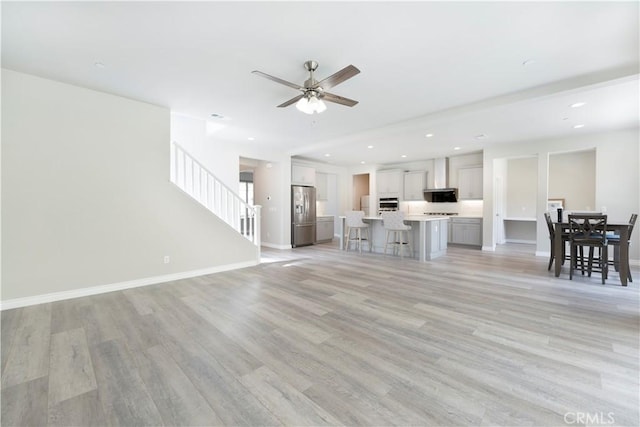 unfurnished living room featuring stairs, baseboards, light wood finished floors, and ceiling fan