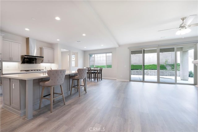 kitchen featuring baseboards, wall chimney range hood, light countertops, an island with sink, and light wood-style floors