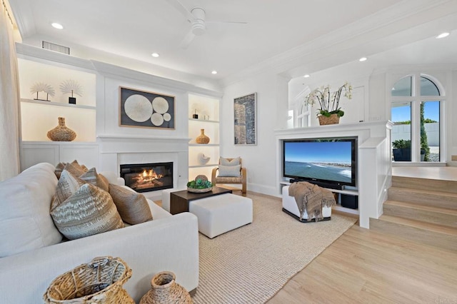 living room featuring built in shelves, ornamental molding, stairs, a glass covered fireplace, and light wood-type flooring