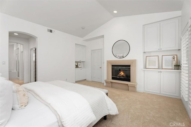 bedroom featuring visible vents, arched walkways, vaulted ceiling, a glass covered fireplace, and light colored carpet