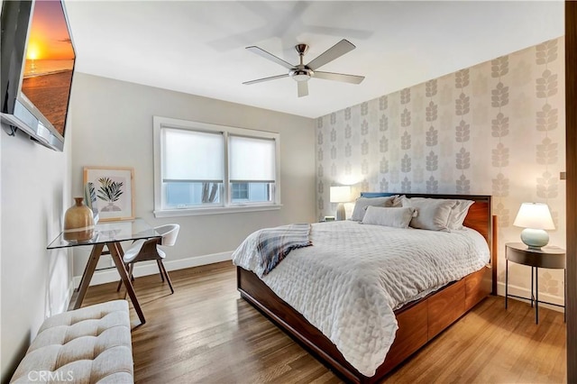 bedroom featuring a ceiling fan, wood finished floors, baseboards, wallpapered walls, and an accent wall