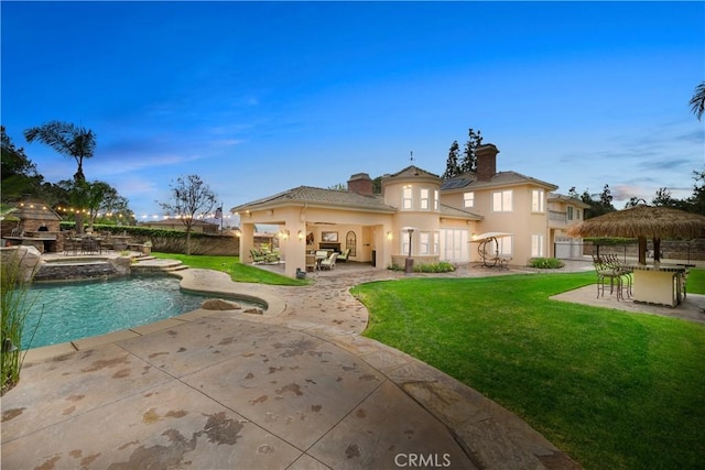 back of property at dusk featuring stucco siding, a fireplace, a chimney, a yard, and a patio