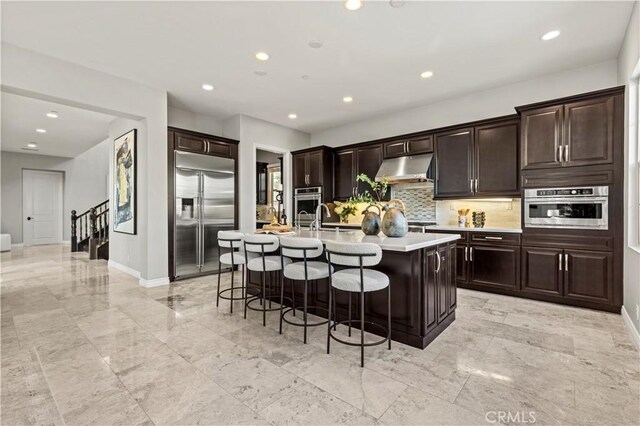 kitchen with a kitchen island with sink, light countertops, appliances with stainless steel finishes, under cabinet range hood, and a kitchen bar