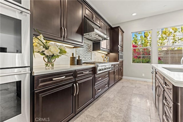 kitchen featuring baseboards, under cabinet range hood, light countertops, decorative backsplash, and appliances with stainless steel finishes