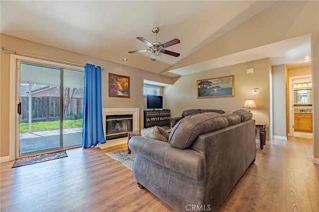 living area with a glass covered fireplace, light wood finished floors, baseboards, ceiling fan, and vaulted ceiling