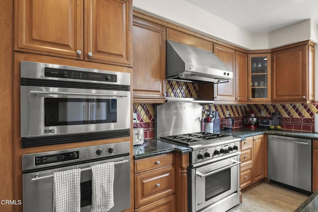 kitchen featuring brown cabinets, dark stone countertops, backsplash, wall chimney range hood, and appliances with stainless steel finishes