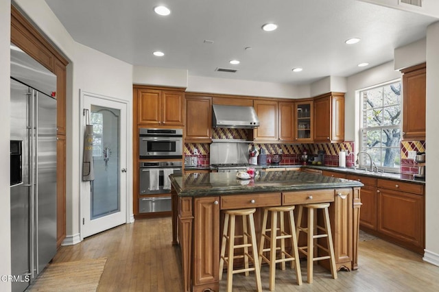 kitchen with brown cabinetry, wall chimney exhaust hood, a warming drawer, and appliances with stainless steel finishes