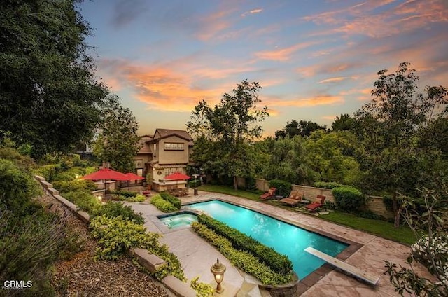 pool at dusk with a patio, a pool with connected hot tub, fence, and a diving board