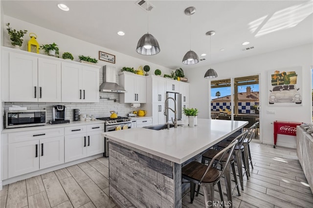 kitchen with visible vents, a kitchen island with sink, a sink, stainless steel appliances, and wall chimney exhaust hood