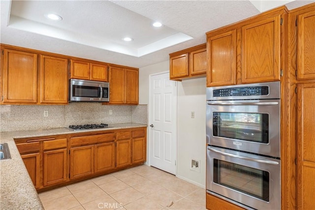 kitchen featuring backsplash, stainless steel appliances, brown cabinetry, light countertops, and a raised ceiling