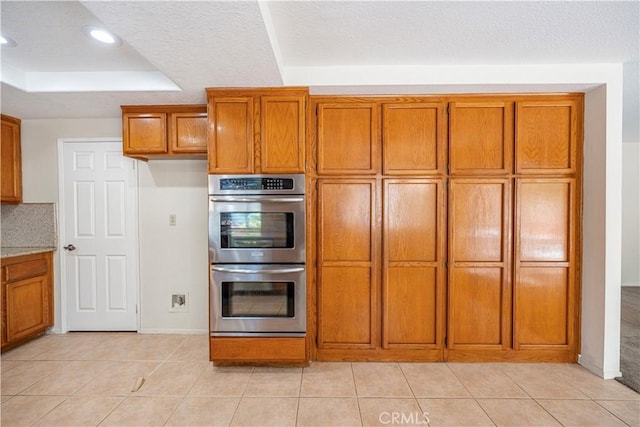 kitchen featuring light tile patterned floors, stainless steel double oven, a textured ceiling, a raised ceiling, and brown cabinets