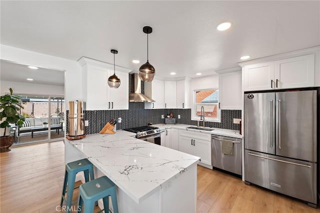 kitchen with a peninsula, a sink, appliances with stainless steel finishes, white cabinetry, and wall chimney range hood