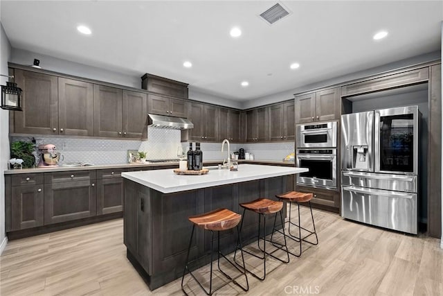 kitchen featuring light wood-style flooring, stainless steel appliances, light countertops, dark brown cabinetry, and under cabinet range hood