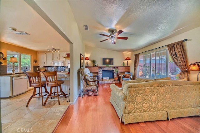 living room featuring visible vents, light wood finished floors, ceiling fan, a textured ceiling, and a brick fireplace