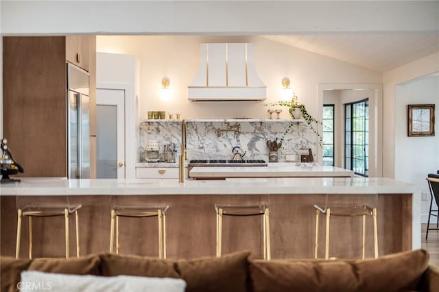 kitchen with backsplash, built in refrigerator, lofted ceiling, custom range hood, and stove