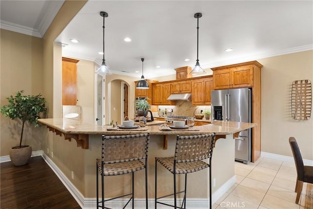 kitchen with arched walkways, high end fridge, ornamental molding, under cabinet range hood, and a kitchen bar