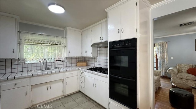 kitchen with under cabinet range hood, tile countertops, ornamental molding, black appliances, and a sink