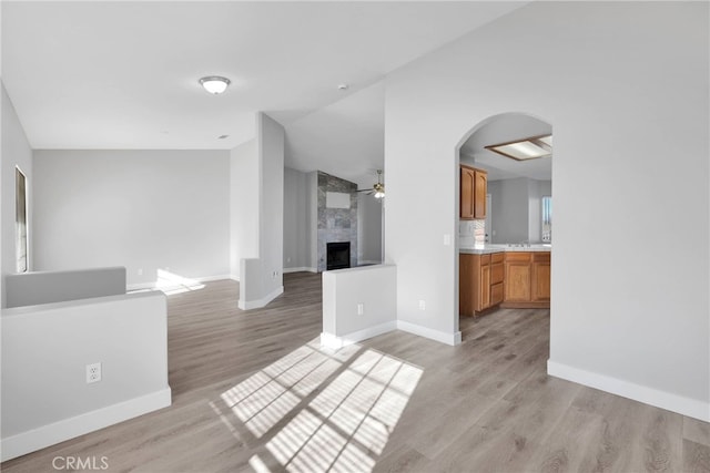 unfurnished living room with baseboards, a fireplace, arched walkways, ceiling fan, and light wood-type flooring