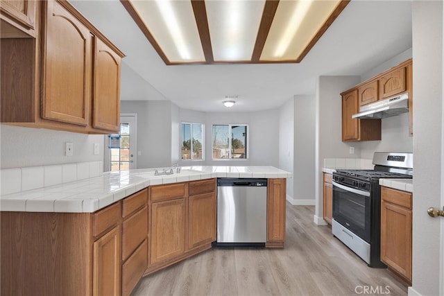 kitchen with under cabinet range hood, tile countertops, light wood-style flooring, appliances with stainless steel finishes, and a peninsula