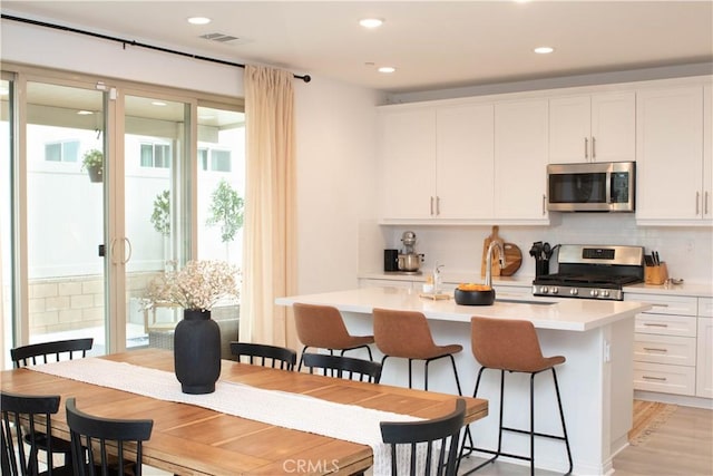 kitchen with a center island with sink, backsplash, white cabinetry, stainless steel appliances, and light countertops