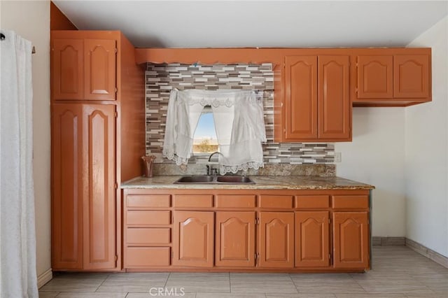 kitchen with a sink, brown cabinetry, decorative backsplash, baseboards, and light stone countertops