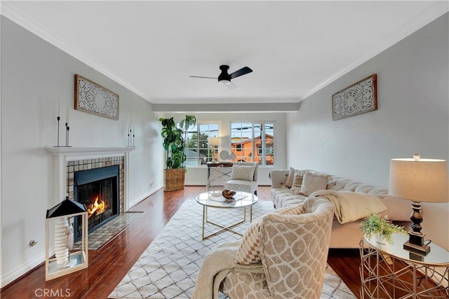 living room featuring wood finished floors, baseboards, ceiling fan, a tile fireplace, and crown molding