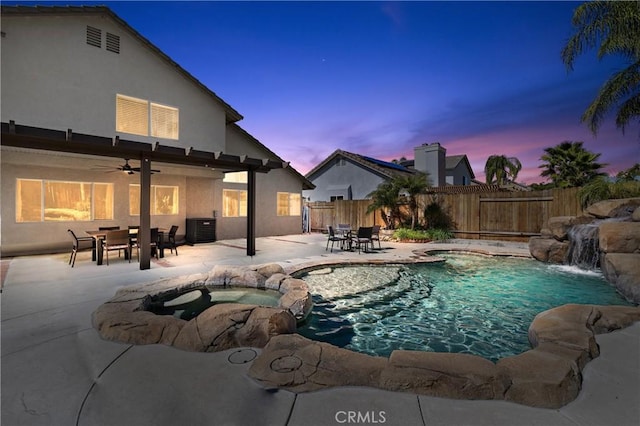 pool at dusk featuring a ceiling fan, a pool with connected hot tub, fence, outdoor dining area, and a patio area