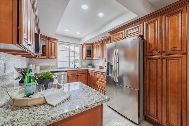 kitchen featuring backsplash, glass insert cabinets, light stone countertops, appliances with stainless steel finishes, and brown cabinetry