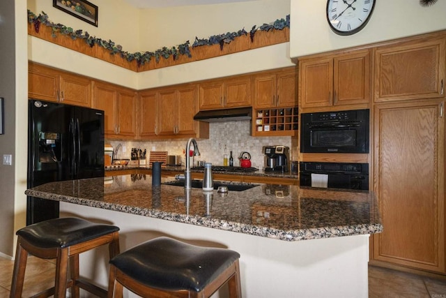 kitchen with tasteful backsplash, under cabinet range hood, dark stone countertops, brown cabinets, and black refrigerator with ice dispenser