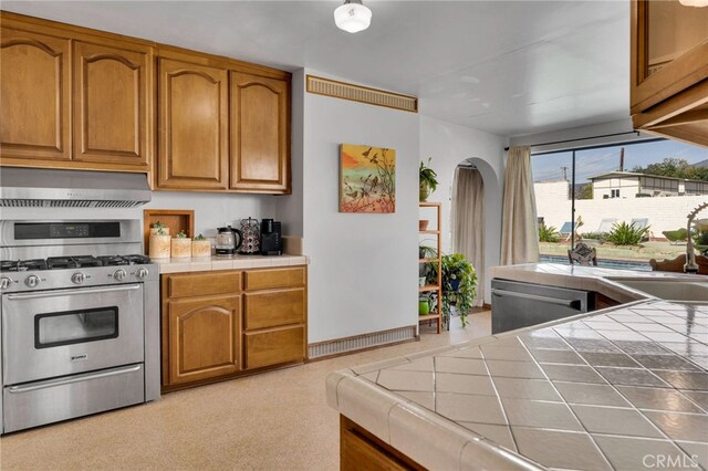 kitchen featuring tile countertops, appliances with stainless steel finishes, exhaust hood, brown cabinetry, and a sink