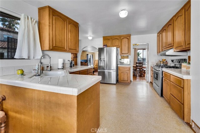 kitchen with under cabinet range hood, a peninsula, brown cabinetry, stainless steel appliances, and a sink