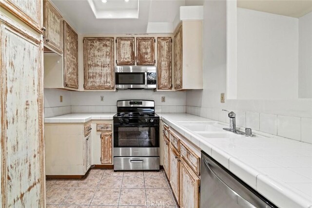 kitchen featuring tile countertops, light tile patterned floors, a sink, stainless steel appliances, and a raised ceiling