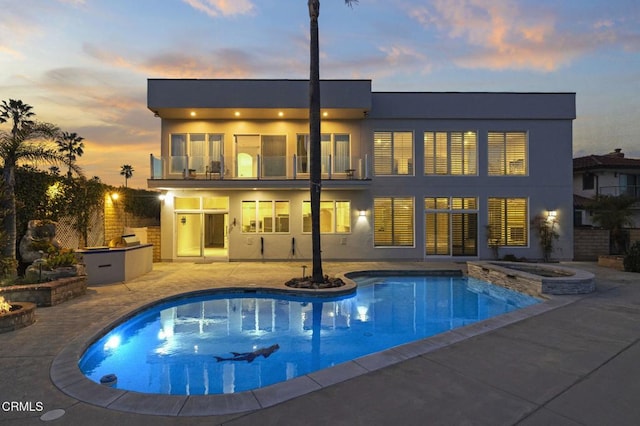 back of house at dusk with a pool with connected hot tub, exterior kitchen, stucco siding, a balcony, and a patio area