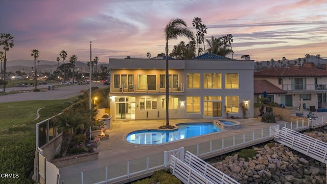 back of property at dusk with a balcony, fence, a pool with connected hot tub, stucco siding, and a patio area