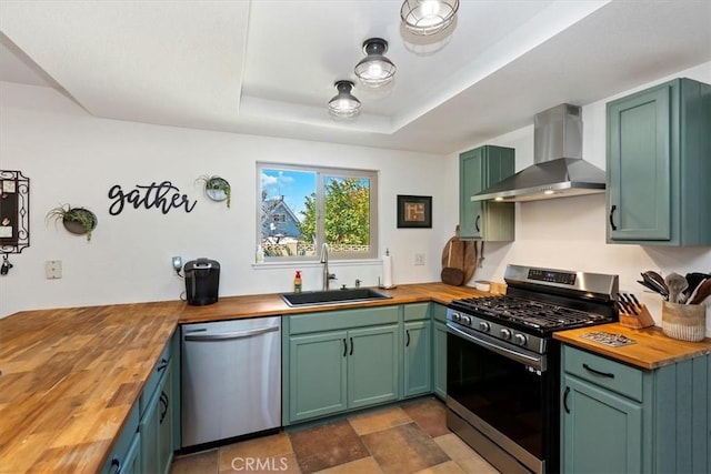 kitchen featuring butcher block countertops, a tray ceiling, appliances with stainless steel finishes, wall chimney exhaust hood, and a sink