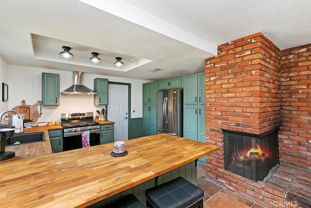 kitchen with a sink, stainless steel appliances, green cabinets, wall chimney range hood, and wooden counters