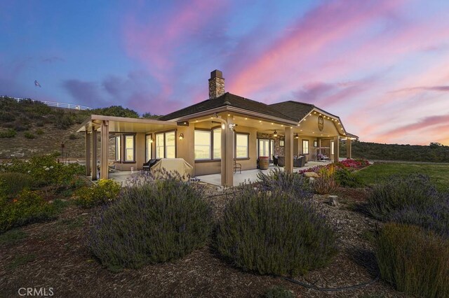 back of house at dusk with stucco siding, a ceiling fan, a chimney, and a patio area